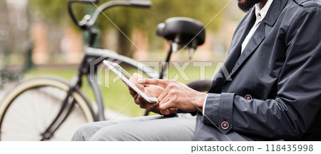 A businessman is sitting on a bench in a park, using a digital tablet with his bicycle close by. The atmosphere is tranquil and efficient, showing a modern work-life balance, cropped 118435998