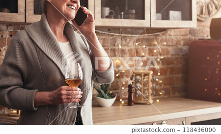 A senior woman is on her phone while holding a glass of wine in her kitchen. She is smiling and appears to be enjoying a conversation, cropped, copy space 118436000
