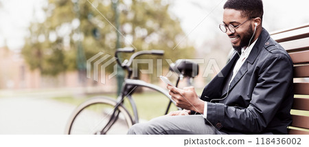 A professional black man is sitting in a park with his phone and bicycle beside him. The mood is contemporary and relaxed, reflecting a modern lifestyle in a natural setting. 118436002