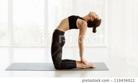 A young woman is shown in a serene yoga studio performing Camel Pose, on a yoga mat. She is dressed in black athletic wear and the daylight from the surrounding windows illuminates the space A young woman is shown in a serene yoga studio performing Camel Pose, on a yoga mat. She is dressed in black athletic wear and the daylight from the surrounding windows illuminates the space 118436022
