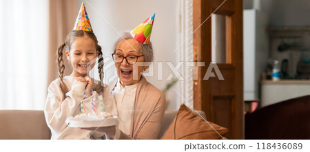 A young girl wearing a party hat proudly presents a birthday cake to her grandmother, who expresses excitement and joy. The cozy living room setting enhances the celebration's warmth. 118436089