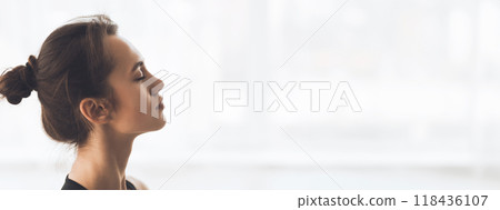 A young woman sits peacefully, eyes closed, in a well-lit room, practicing mindfulness and meditation, copy space 118436107