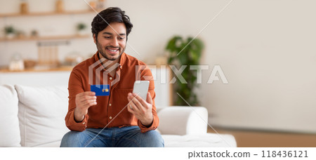 Indian man seated comfortably on a sofa is smiling as he compares details on his mobile phone while holding a credit card. The bright indoor setting creates a relaxed atmosphere. 118436121