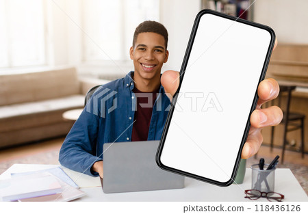 African American young man sits at a desk in a well-lit room, working on a laptop. He holds up a smartphone, smiling cheerfully, ready to share information or a message. African American young man sits at a desk in a well-lit room, working on a laptop. He holds up a smartphone, smiling cheerfully, ready to share information or a message. 118436126