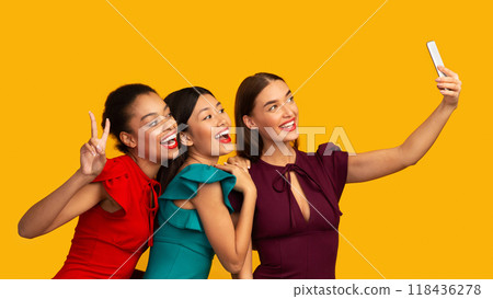 Party Selfie. Three Millennial Girls Using Phone Taking Pictures Of Themselves Having Fun Over Yellow Background. Studio Shot, Panorama 118436278