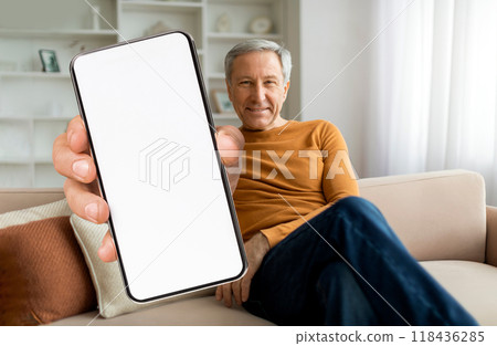 An older man relaxes on a couch in a well-lit room, smiling at the camera while holding up a blank smartphone. The setting suggests comfort and modern technology. 118436285