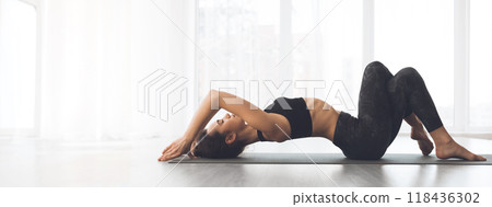A woman performs a yoga pose on her mat in a well-lit studio, focusing on her breath and relaxation, copy space 118436302