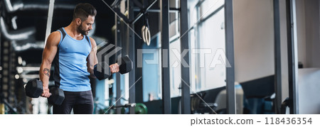 A man in a blue tank top is performing bicep curls with dumbbells in a well-lit gym, copy space A man in a blue tank top is performing bicep curls with dumbbells in a well-lit gym, copy space 118436354