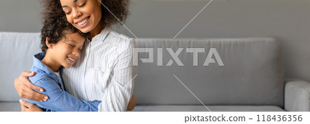 African American mother smiles brightly as she embraces her son, both feeling a sense of love and warmth in their cozy living room during a sunny afternoon. African American mother smiles brightly as she embraces her son, both feeling a sense of love and warmth in their cozy living room during a sunny afternoon. 118436356