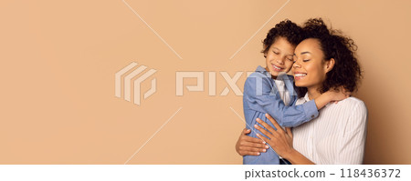 In a warm, cozy moment, African American mother holds her young child closely, both smiling with joy. The soft background emphasizes their connection and love for one another. 118436372