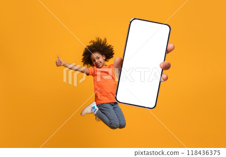 A young black child in an orange shirt enthusiastically jumps in the air while giving a thumbs up, holding a large blank phone with a vibrant yellow background that enhances the cheerful atmosphere. A young black child in an orange shirt enthusiastically jumps in the air while giving a thumbs up, holding a large blank phone with a vibrant yellow background that enhances the cheerful atmosphere. 118436375