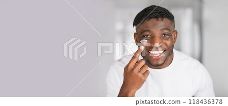 A cheerful young African American man appears in a well-lit bathroom, applying a skincare cream to his face. He smiles confidently, showcasing a positive self-care routine. 118436378