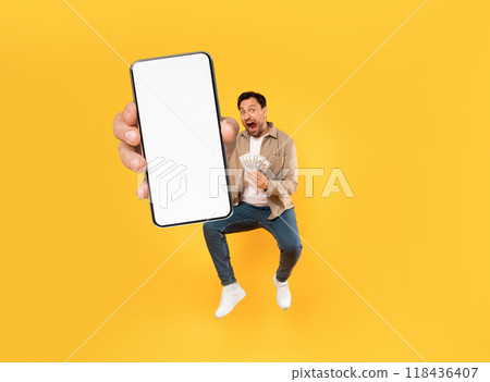 A man leaps in the air while holding a handful of money in one hand and a smartphone displaying a blank screen in the other, set against a vibrant yellow backdrop, showcasing enthusiasm and surprise. 118436407