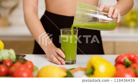 A woman in a black skirt pours a green smoothie into a glass from a pitcher, with fresh produce visible on the counter. 118436439