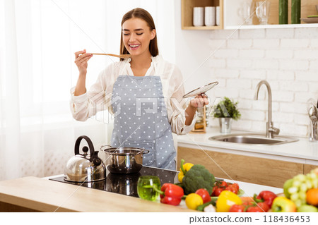 A happy woman in an apron tastes food while cooking in her kitchen. She is surrounded by fresh vegetables and cooking supplies. A happy woman in an apron tastes food while cooking in her kitchen. She is surrounded by fresh vegetables and cooking supplies. 118436453