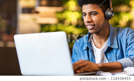Closeup of African American young guy wearing headphones is focused as he works on a laptop. His hands are typing on the keyboard, and the laptop screen displays graphs and data. Closeup of African American young guy wearing headphones is focused as he works on a laptop. His hands are typing on the keyboard, and the laptop screen displays graphs and data. 118436455