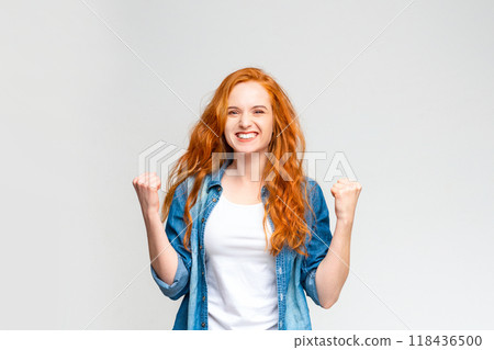 A woman with long red hair is exuberant, raising her clenched fists in a gesture of victory or celebration, wearing a casual white t-shirt and a blue denim shirt against a neutral grey backdrop. A woman with long red hair is exuberant, raising her clenched fists in a gesture of victory or celebration, wearing a casual white t-shirt and a blue denim shirt against a neutral grey backdrop. 118436500