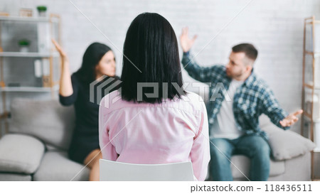 A woman sits on a chair, facing away from the camera, as a couple argues on a couch in the background. The woman is likely a therapist, witnessing the couples conflict during a session. 118436511
