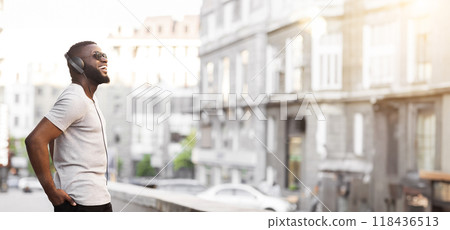 A man with headphones on is smiling brightly as he walks through a city street. He is wearing sunglasses and a white t-shirt. A man with headphones on is smiling brightly as he walks through a city street. He is wearing sunglasses and a white t-shirt. 118436513