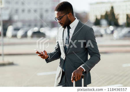 African American man in a suit is using his phone while standing with luggage near an airport entrance. The setting is modern and professional, highlighting business travel. 118436557