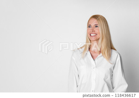 A smiling woman with blonde hair and wearing a white collared shirt stands in front of a white background and looks off-camera. She appears to be in her 40s or 50s, copy space 118436617