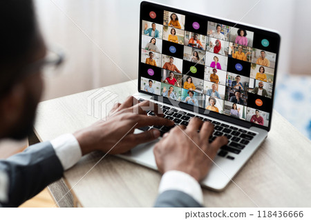 Black man in a grey suit and white shirt sits at a desk using a laptop to participate in a video conference. His hands are on the keyboard and the laptop screen shows participants in a video chat 118436666