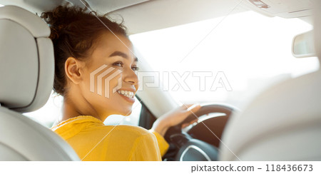 African American young woman with curly brown hair is driving a car. She is smiling and looking out the window. The sun is shining brightly, and the woman is wearing a yellow shirt. 118436673