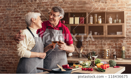 An older couple, both wearing aprons, are standing in their kitchen, smiling and toasting with glasses of wine. The kitchen has brick walls and wooden cabinets. 118436675
