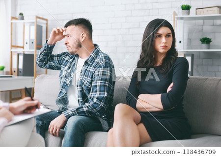 A man and a woman sit on a couch in a therapy session. The man is looking down, with his hand on his forehead, while the woman is sitting with her arms crossed, looking straight ahead 118436709