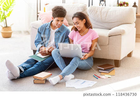 African American teen couple sitting on the floor in their home, studying together. The girl is using a laptop while the guy is taking notes in a notebook. 118436730