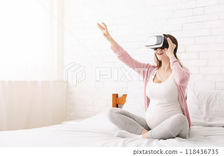 Expecting woman is sitting on a bed, wearing a virtual reality headset. She appears engaged in the virtual experience, with a focused expression on her face, copy space 118436735