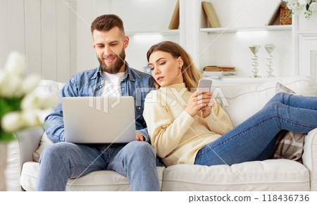 A couple sits comfortably on a white sofa, the man using a laptop while the woman holds a smartphone, in a bright, modern living room. 118436736