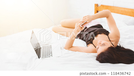 A woman in a black nightgown is lying on a bed, using her smartphone while a laptop sits nearby on white, neatly arranged sheets. Sunlight brightly illuminates the room 118436742