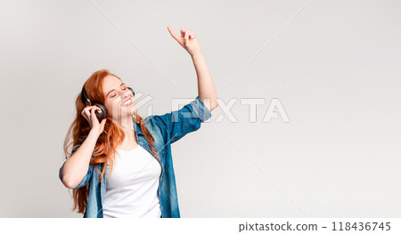 A young woman with red hair, wearing casual denim jacket over a white top, is captured mid-dance with radiant smile. She is lost in the music, enjoying the rhythm through her headphones, copy space 118436745