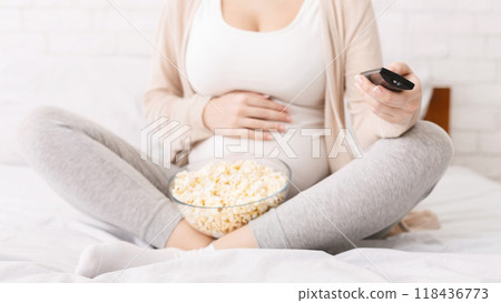 Cropped of pregnant woman is seen sitting on a bed, holding a bowl of popcorn and a remote control. She seems relaxed and comfortable in the scene. Cropped of pregnant woman is seen sitting on a bed, holding a bowl of popcorn and a remote control. She seems relaxed and comfortable in the scene. 118436773
