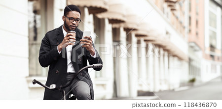 An African American businessman is biking to the office while using his phone. The scene is energetic and urban, highlighting the blend of modern technology and eco-friendly commuting. 118436870