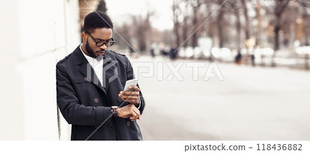 Black businessman walking to office by street, holding phone, checking time, looking at watch, panorama with copy space Black businessman walking to office by street, holding phone, checking time, looking at watch, panorama with copy space 118436882