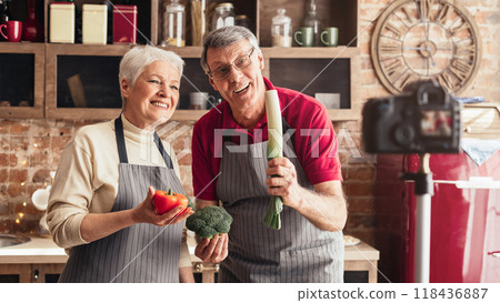 A senior couple is filming a cooking show in their home kitchen. The woman is holding a red cabbage while the man is gesturing with his hand and smiling at the camera. 118436887
