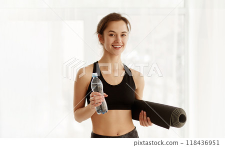 A woman stands holding a water bottle and a yoga mat in an indoor setting. She appears ready for a workout or yoga session. 118436951