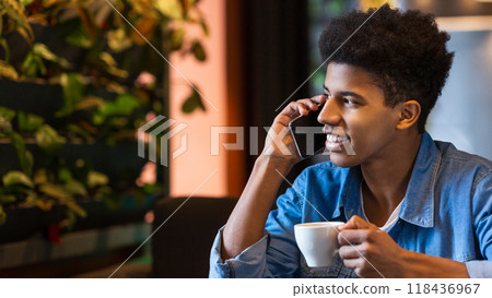 African American guy is seated at a table with a cup of coffee in front of him, engaged in a phone conversation. He appears focused and attentive to the call. African American guy is seated at a table with a cup of coffee in front of him, engaged in a phone conversation. He appears focused and attentive to the call. 118436967