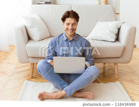 A man is sitting on the floor in a living room, using a laptop. He is smiling and appears to be relaxed. 118436990