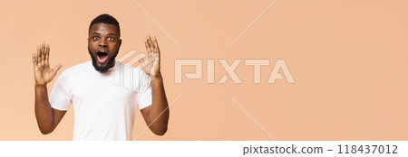 African American young man with short hair and a white t-shirt looks surprised with his mouth open and hands raised in front of a beige background, panorama with copy space 118437012
