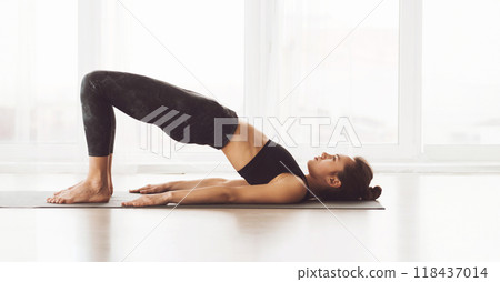 A woman is performing a yoga bridge pose on a mat in a well-lit studio with large windows. She is focused and appears to be in the middle of a workout session. A woman is performing a yoga bridge pose on a mat in a well-lit studio with large windows. She is focused and appears to be in the middle of a workout session. 118437014