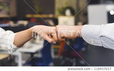 A close-up shows two individuals engaging in a fist bump, a gesture of affirmation or greeting, with one wearing a striped shirt. The background suggests an informal office environment, cropped 118437017