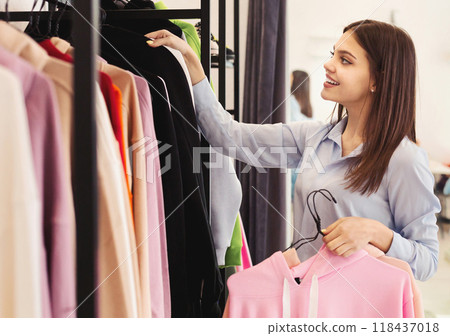 A woman browsing through a rack of clothes in a store, examining different items with interest and possibly trying some on. She is standing alone, focused on the garments in front of her. 118437018