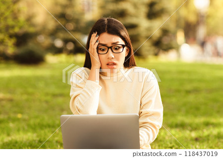 Asian young woman dressed in a cream sweater sits on the grass in a sunny park, looking surprised and alarmed as she stares at her laptop screen. Asian young woman dressed in a cream sweater sits on the grass in a sunny park, looking surprised and alarmed as she stares at her laptop screen. 118437019