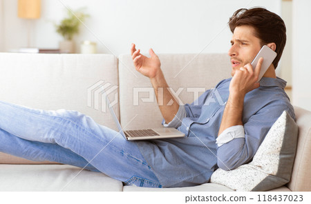 A man sits on a couch, talking on his phone while working on his laptop. A man sits on a couch, talking on his phone while working on his laptop. 118437023