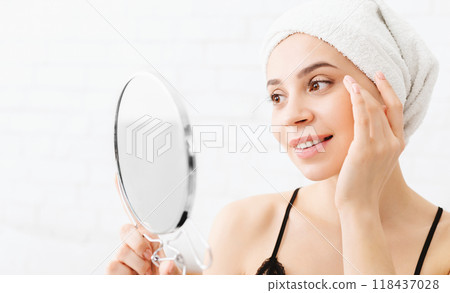 A woman with a towel wrapped around her head is examining her face in a mirror after morning shower, closeup A woman with a towel wrapped around her head is examining her face in a mirror after morning shower, closeup 118437028