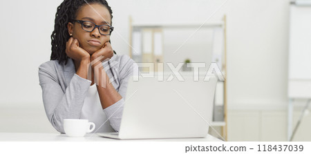 A tired black woman is using her laptop at an office desk. The mood is dull and weary, with a cluttered desk and uninspired surroundings, showing her lack of engagement with her work. A tired black woman is using her laptop at an office desk. The mood is dull and weary, with a cluttered desk and uninspired surroundings, showing her lack of engagement with her work. 118437039