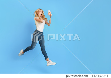 A young woman with blonde hair is jumping in the air with her arms outstretched. She is looking at her phone in surprise, as if she has just received good news, copy space 118437051